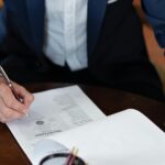 Person unterzeichnet Handwerkervertrag oder Sanierungsdokumentation am Tisch mit Zeugen