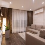 Modernes Wohnzimmer mit Klimaanlage an der Wand, beigem Sofa, braunen Vorhängen und Holzboden nach Sanierungsumbau
