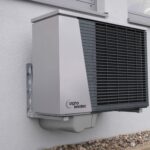Moderne Wärmepumpe an Hausfassade montiert, weiße Außeneinheit mit Kühlergitter und Kondensatablauf für Sanierung