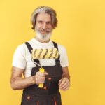 Handwerker mit Bart und Malerschürze hält Farbwalze vor gelbem Hintergrund