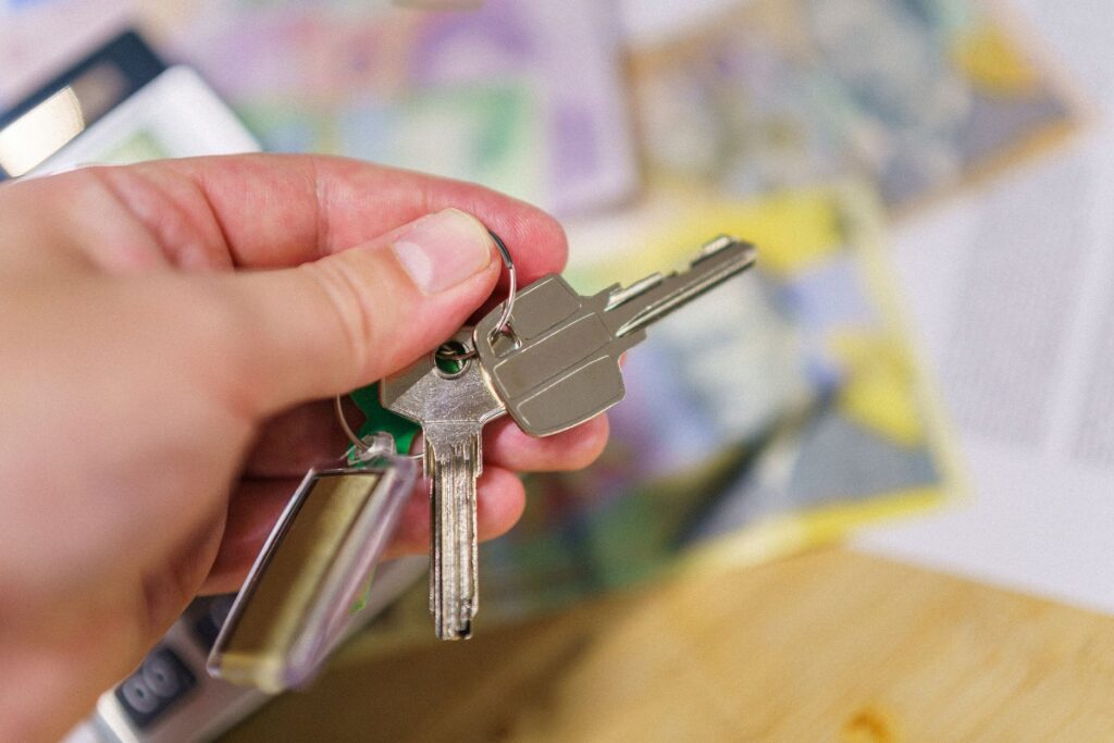 Hand hält Schlüsselpaar mit grünem Kunststoffgriff vor unscharfem Hintergrund mit Unterlagen