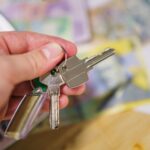 Hand hält Schlüsselpaar mit grünem Kunststoffgriff vor unscharfem Hintergrund mit Unterlagen