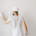 Handwerker mit Schutzhelm und Atemmaske beim Innenwand verputzen trägt weiße Spachtelmasse auf heller Wand auf