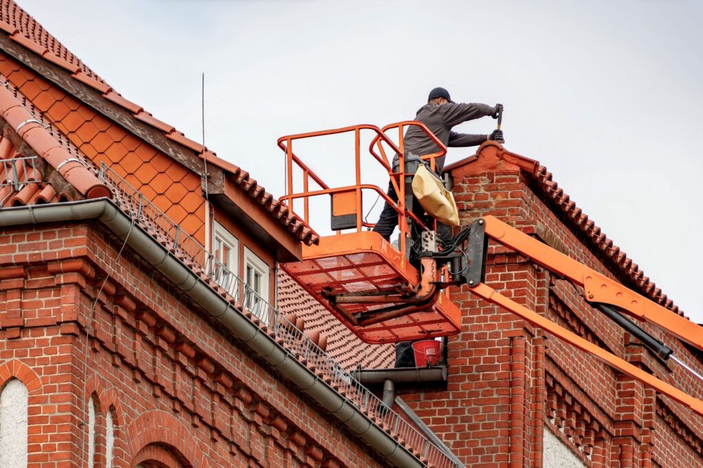 Handwerker auf Hubarbeitsbühne reinigt Dachrinne am Ziegeldach eines Backsteinhauses