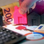 Hand platziert pinkes Hausmodell zwischen schwarzen Häusern auf Finanzunterlagen mit Geldschein und Taschenrechner zur Sanierungsfinanzierung
