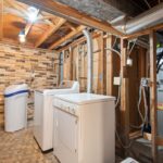Keller-Installationsraum mit Wasserenthärtungsanlage, Waschmaschine und Rohrleitungen unter Holzdecke