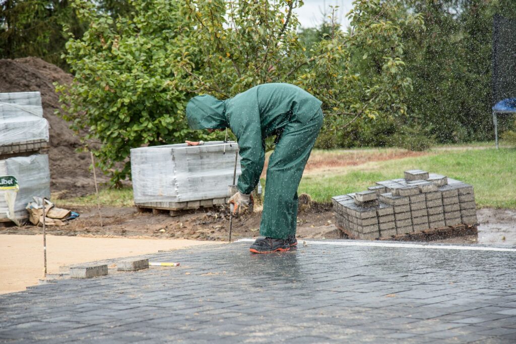 Handwerker verlegt Pflastersteine im Regen auf einer Baustelle mit Materialien im Hintergrund